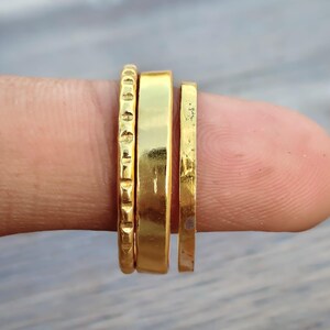 May include: A set of three gold-toned rings featuring distinct textures. The first ring has a repeating notched pattern, the second is a wide smooth band, and the third is a thin, narrow band with a slightly hammered or irregular surface finish.