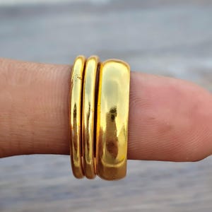 May include: A set of three gold-coloured rings with a polished, reflective finish. The set consists of two thin, rounded bands and one wider, flat-profile band. The rings are stacked together, showcasing a smooth, metallic surface and a bright yellow-gold tone.
