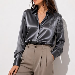 May include: A long-sleeved charcoal gray satin button-down shirt with a classic collar and wide cuffs, paired with high-waisted beige trousers featuring front pleats. The shirt has a smooth, reflective finish and a hidden button placket design.