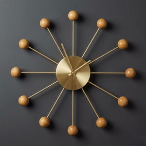 May include: A sunburst wall clock featuring a central circular brushed brass face with matching thin metal spokes radiating outward. Each spoke is tipped with a smooth, round, natural light-toned wooden ball, and the clock includes two simple brass hands.