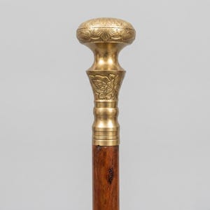 May include: A walking stick featuring a polished brass handle with an ornate, engraved floral pattern on the rounded top and the tapered neck. The handle is securely attached to a dark brown wooden shaft with a visible natural wood grain texture.