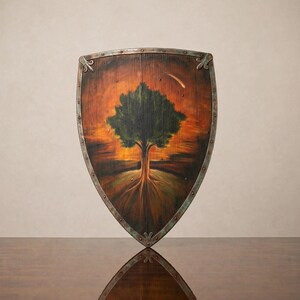 May include: A wooden shield featuring a painted green tree against a vibrant orange and brown sunset background. The shield has a dark metal border with decorative rivets and metal corner accents, showcasing a weathered texture and visible wood grain.