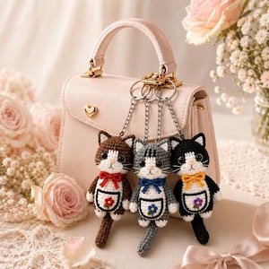 May include: A light pink handbag featuring three crocheted cat keychains attached to a silver metal ring. Each cat has a white belly patch with a small flower, a colored bow tie, and a long tail. The cats are brown, gray, and black.