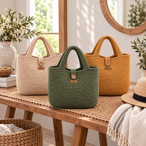 Puede incluir: Tres bolsos de ganchillo en color beige, verde oliva y amarillo mostaza. Cada bolso cuenta con dos asas superiores, un cierre de botón de madera en el centro y una pequeña etiqueta rectangular de cuero con un icono de hoja en el frente.