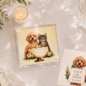 Puede incluir: Una placa acrílica cuadrada con la ilustración de un cachorro dorado y un gato atigrado sentados tras un pastel blanco decorado con rosas color melocotón. Al lado, una tarjeta muestra a un cachorro sosteniendo un cartel que dice SAVE the DATE 06.14.2026.