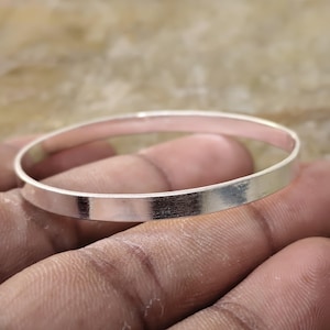 May include: A simple, polished silver-toned bangle bracelet with a smooth, flat outer surface and a circular shape. The metal band has a reflective finish that catches the light, showcasing a clean, minimalist design without any added embellishments.
