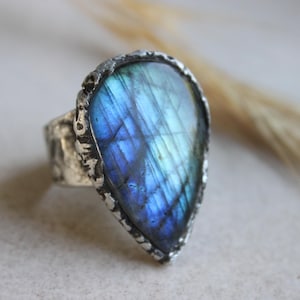 Op de afbeelding: Een druppelvormige labradoriet ring met een levendige blauwe en paarse iriserende glans. De steen is gezet in een getextureerde, donker zilverkleurige metalen band met een ruwe, onregelmatige rand die het gepolijste oppervlak van de edelsteen omlijst.