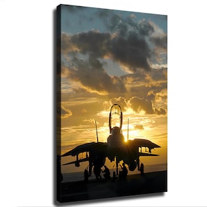 May include: A fighter jet silhouette positioned on a flight deck during a golden sunset. The aircraft features an open cockpit canopy and extended wings, with several crew members visible near the landing gear against the bright horizon.