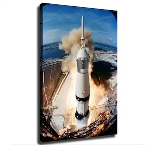 May include: A tall white and black rocket stands vertically on a launch pad, emitting a large plume of tan smoke from its base. The scene is captured from a high-angle perspective, showing the surrounding landscape and blue sky above.