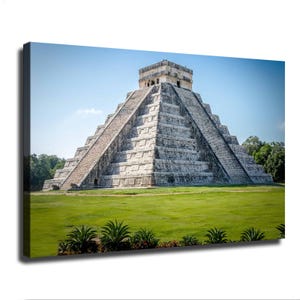 May include: A large, gray stone pyramid with a stepped design and a flat top structure, situated on a vibrant green lawn under a clear blue sky. The ancient monument features symmetrical staircases leading to the summit, surrounded by trees.