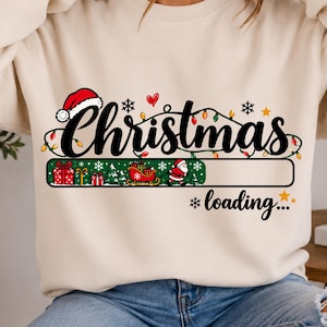 May include: A cream-colored crewneck sweatshirt featuring a graphic print with the text "Christmas loading..." in black script. The design includes a progress bar with gift boxes, a sleigh, and Santa Claus, accented by colorful string lights.