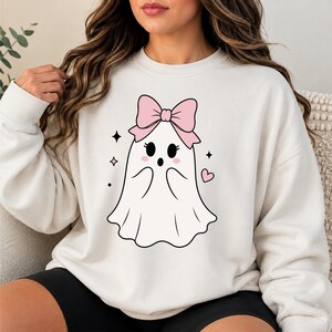 May include: A cream-colored crewneck sweatshirt featuring a central graphic of a white ghost with a large pink bow on its head, black eyes, a small mouth, pink cheeks, and surrounding small black star and heart icons printed on the front.