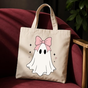 May include: A beige canvas tote bag featuring a central graphic of a white ghost with black oval eyes. The ghost is adorned with a large, light pink bow on its head and is surrounded by small black starburst icons and tiny pink heart shapes.