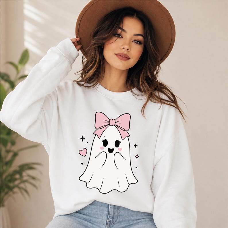 May include: A white long-sleeve sweatshirt featuring a central graphic of a white ghost with a large pink bow on its head, pink cheeks, and black eyes. The design includes small pink hearts and black star-shaped sparkles surrounding the ghost.