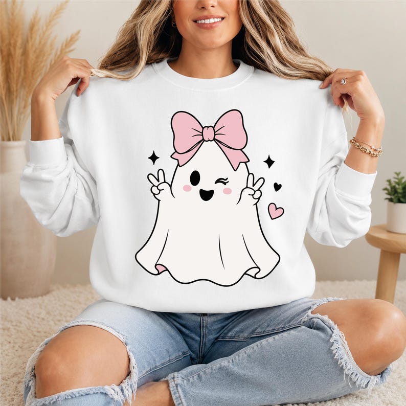 May include: A white long-sleeve sweatshirt featuring a central graphic of a white ghost with a large pink bow on its head. The ghost is winking, smiling, and making peace signs with both hands, surrounded by small black stars and pink hearts.