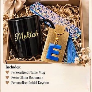 May include: A gift box containing a black ceramic mug with the name Mehtab in gold script, a blue resin initial E keychain on a gold card, and a floral patterned resin bookmark with a blue tassel, all nestled in shredded paper filler.