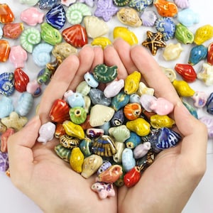 May include: A collection of small, glazed ceramic beads in various shapes, including seashells, starfish, and fish. The beads feature a vibrant palette of red, blue, green, yellow, pink, and white, with glossy finishes and visible holes for threading.