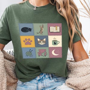 Puede incluir: Una camiseta de manga corta color verde musgo con un gráfico central de cuadrícula compuesto por nueve cuadrados. Los cuadrados muestran ilustraciones de un gato durmiendo, un gato de pie, un libro abierto, una huella, una cara de gato, una taza, lana, un sillón y una luna.