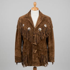May include: A brown suede jacket featuring long fringe detailing across the chest and pockets. The garment includes silver-toned circular metal concho accents along the upper chest and secures with wooden toggle buttons down the center front.