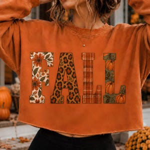 Könnte beinhalten: Ein orangefarbenes Langarm-Sweatshirt mit dem Wort FALL in großen Blockbuchstaben. Der Buchstabe F hat ein Blumenmuster, A ein Leopardenmuster, L ein Karomuster und L eine Grafik mit Kürbissen und Ranken. Das Sweatshirt hat einen offenen Saum.