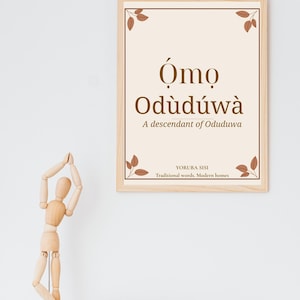 May include: A framed wall art print featuring the text Ómọ Odùdúwà and A descendant of Oduduwa in brown font on a cream background. The print is enclosed in a light wood frame and decorated with small brown leaf illustrations in each corner.