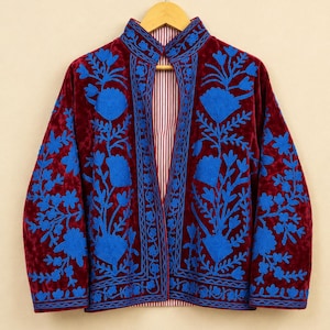 Könnte beinhalten: Eine tiefburgunderrote Samtjacke mit leuchtend blauer Blumenstickerei auf den Vorderteilen und dem Kragen. Das Kleidungsstück hat einen hohen Ausschnitt mit einer zentralen vertikalen Öffnung und ein rot-weiß gestreiftes Innenfutter. Text: HEVENCRAFTINDIA.