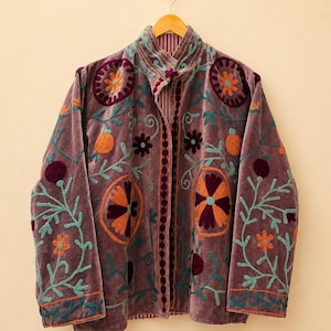 Könnte beinhalten: Eine mauvefarbene Samtjacke mit Stehkragen und langen Ärmeln. Das Kleidungsstück ist mit gestickten Blumen- und Kreismotiven in Orange, Petrol und Dunkellila verziert. Der Text HEVENCRAFTINDIA ist oben rechts im Bild zu sehen.