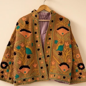 May include: A tan jacket featuring vibrant floral embroidery in shades of orange, teal, yellow, and black. The garment has a mandarin collar, long sleeves, and an open front design. The interior lining is visible and displays a purple and white striped pattern. The text HEVENCRAFT INDIA is printed in the upper right corner.