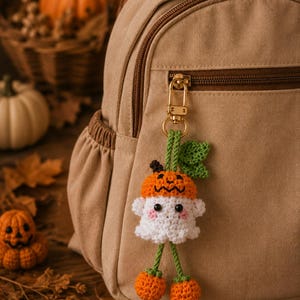 Könnte beinhalten: Ein gehäkelter Schlüsselanhänger in Form eines weißen Geistes mit einem orangefarbenen Kürbishut und einem grünen Blatt, befestigt an einem goldenen Metallkarabiner an einem beigen Rucksack, mit zwei kleinen orangefarbenen Kürbissen darunter.