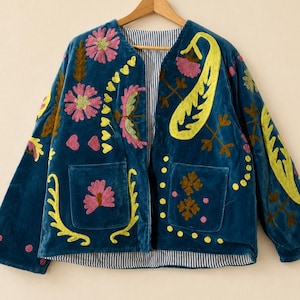 Könnte beinhalten: Eine petrolfarbene Samtjacke mit lebendigen Blumen- und Paisley-Stickereien in Pink und Gelb. Das offene Kleidungsstück verfügt über zwei Vordertaschen und ein blau-weiß gestreiftes Innenfutter. Der Text HEVENCRAFTINDIA ist oben rechts zu sehen.