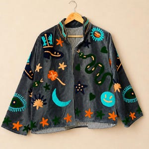 Könnte beinhalten: Eine anthrazitfarbene Samtjacke mit bunten Stickmotiven, darunter eine grüne Schlange, ein blauer Halbmond, ein Cowboystiefel, ein Smiley-Gesicht und verschiedene Sterne. Die Jacke hat einen Stehkragen und ein schwarz-weiß gestreiftes Innenfutter.