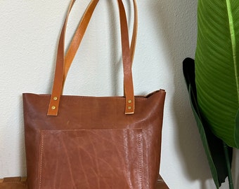 May include: A brown leather tote bag with two straps and a front pocket. The bag is sitting on a woven brown surface.