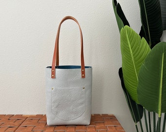May include: A white leather tote bag with brown leather handles and a small pocket on the front. The bag has a subtle embossed floral pattern.