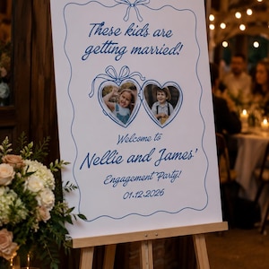 Puede incluir: Un letrero rectangular blanco sobre un caballete de madera con texto azul y un borde azul con un lazo. El texto dice: These kids are getting married! Welcome to Nellie and James' Engagement Party! 01.12.2026. Dos marcos de fotos en forma de corazón muestran a una niña y un niño.