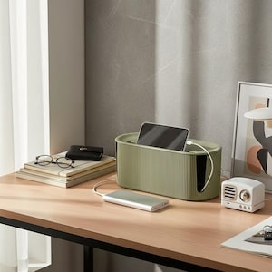 Könnte beinhalten: Eine olivgrüne Kabelmanagement-Box mit gerippter Außenstruktur und rechteckiger Form. Die Box verfügt über einen Schlitz an der Oberseite für ein schwarzes Smartphone und eine seitliche Öffnung für Kabel, mit einer silbernen Powerbank, die über ein weißes Kabel verbunden ist.