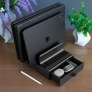 May include: A black desktop organiser stand featuring vertical slots for holding multiple tablets or laptops, a front ledge for device support, and a small pull-out storage drawer containing a white earbud case and a silver car key fob.