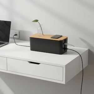 May include: A rectangular black cable management box with a light wood grain lid. The box sits on a white desk and holds a small glass vase with a green leaf and a smartphone. Black power cords extend from the sides of the storage container.