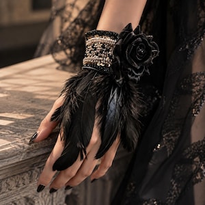 May include: A black wrist cuff featuring a wide band adorned with rows of metallic beads and sequins, topped with a large black fabric rose. The cuff is embellished with long, dark black feathers that extend downwards over the back of the hand.