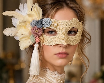 Puede incluir: Un antifaz de encaje dorado para baile de máscaras con un grupo de plumas color crema, pequeñas flores de tela en tonos azul, rosa y verde, perlas blancas y una larga borla color crema que cuelga del lateral del marco que cubre los ojos.