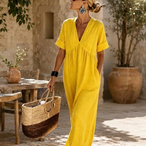 May include: A bright yellow maxi dress with a V-neckline, short cuffed sleeves, and side pockets. The dress is paired with a two-toned woven straw tote bag, black sunglasses, large patterned circular earrings, and a black stacked bangle bracelet.