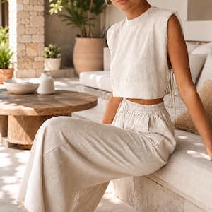 May include: A beige two-piece set consisting of a sleeveless crop top with padded shoulders and side tie closures, paired with high-waisted wide-leg trousers featuring an elasticated waistband with a drawstring and side pockets.