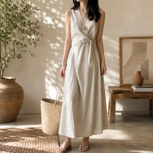 May include: A sleeveless, floor-length wrap dress in a light beige color, featuring a V-neckline and a fabric tie at the waist. The dress is made of a textured material and is paired with a woven straw tote bag and simple flat sandals.