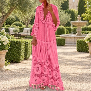 May include: A bright pink maxi dress featuring a deep V-neckline, long sleeves, and a tiered skirt. The garment is adorned with intricate lace trim, decorative tassels along the front opening, and a wide floral lace panel at the hemline.
