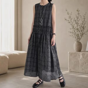 May include: A sleeveless, ankle-length dress in a dark grey and black plaid pattern. The garment features a round neckline with a button placket, a gathered elastic waistline, and a full, flowing skirt. A long silver chain necklace is worn.