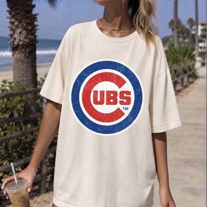Puede incluir: Una camiseta de manga corta color crema con un gráfico circular grande en el pecho. El gráfico muestra un anillo azul desgastado que rodea una letra C roja con el texto CUBS TM en letras blancas de bloque centradas dentro de la C roja.
