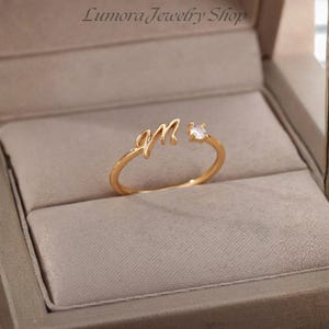 Peut inclure: Une bague ouverte dorée présentant une lettre M en écriture cursive d'un côté et une petite pierre précieuse ronde et transparente sertie de griffes de l'autre. La bague est présentée dans une boîte à bijoux beige avec le texte Lumora Jewelry Shop.