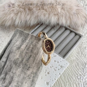 Può includere: Un orologio rigido color oro con cassa ovale e quadrante marrone scuro. L'orologio presenta lancette e indici sottili color oro. Il nome del marchio Naidu è stampato in piccole lettere dorate al centro del quadrante, racchiuso in una cornice di metallo color oro lucido.
