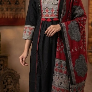 May include: A black three-piece outfit consisting of a long-sleeved tunic with a V-neckline, matching trousers, and a red and black patterned shawl. The tunic features intricate white and red embroidery on the chest and cuffs with small red tassels.