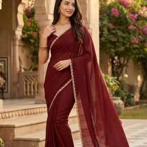 May include: A deep maroon saree featuring a subtle horizontal striped texture throughout the fabric. The garment is finished with a delicate, scalloped gold lace border along the edges, providing a refined contrast to the dark red tone.