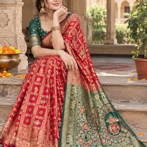 Op de afbeelding: Een rode sari met een goudkleurig geometrisch patroon en een brede groene rand met ingewikkelde gouden motieven, waaronder olifanten en een centraal urnontwerp. Het kledingstuk is gecombineerd met een donkergroene blouse met gouden details op de mouwen.
